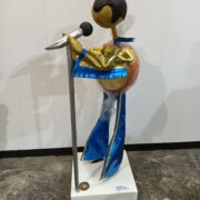 Scultura con performer in pelli iridescenti oro e blu davanti al microfono, opera di PierAngelo Bertolo