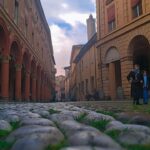Strada acciottolata ripresa dal basso tra edifici storici porticati, con alcuni passanti sullo sfondo e cielo nuvoloso.