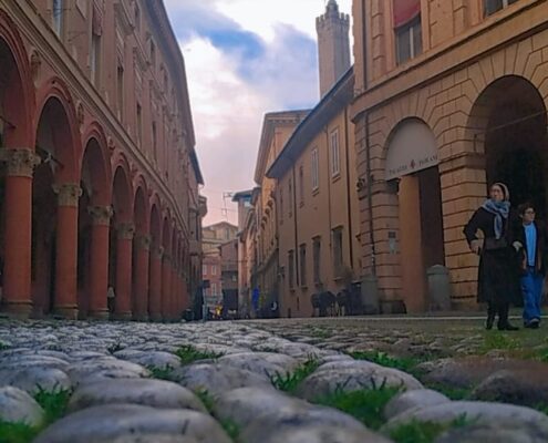 Strada acciottolata ripresa dal basso tra edifici storici porticati, con alcuni passanti sullo sfondo e cielo nuvoloso.