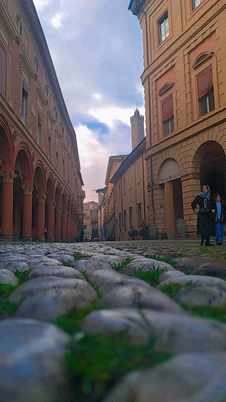 Strada acciottolata ripresa dal basso tra edifici storici porticati, con alcuni passanti sullo sfondo e cielo nuvoloso.