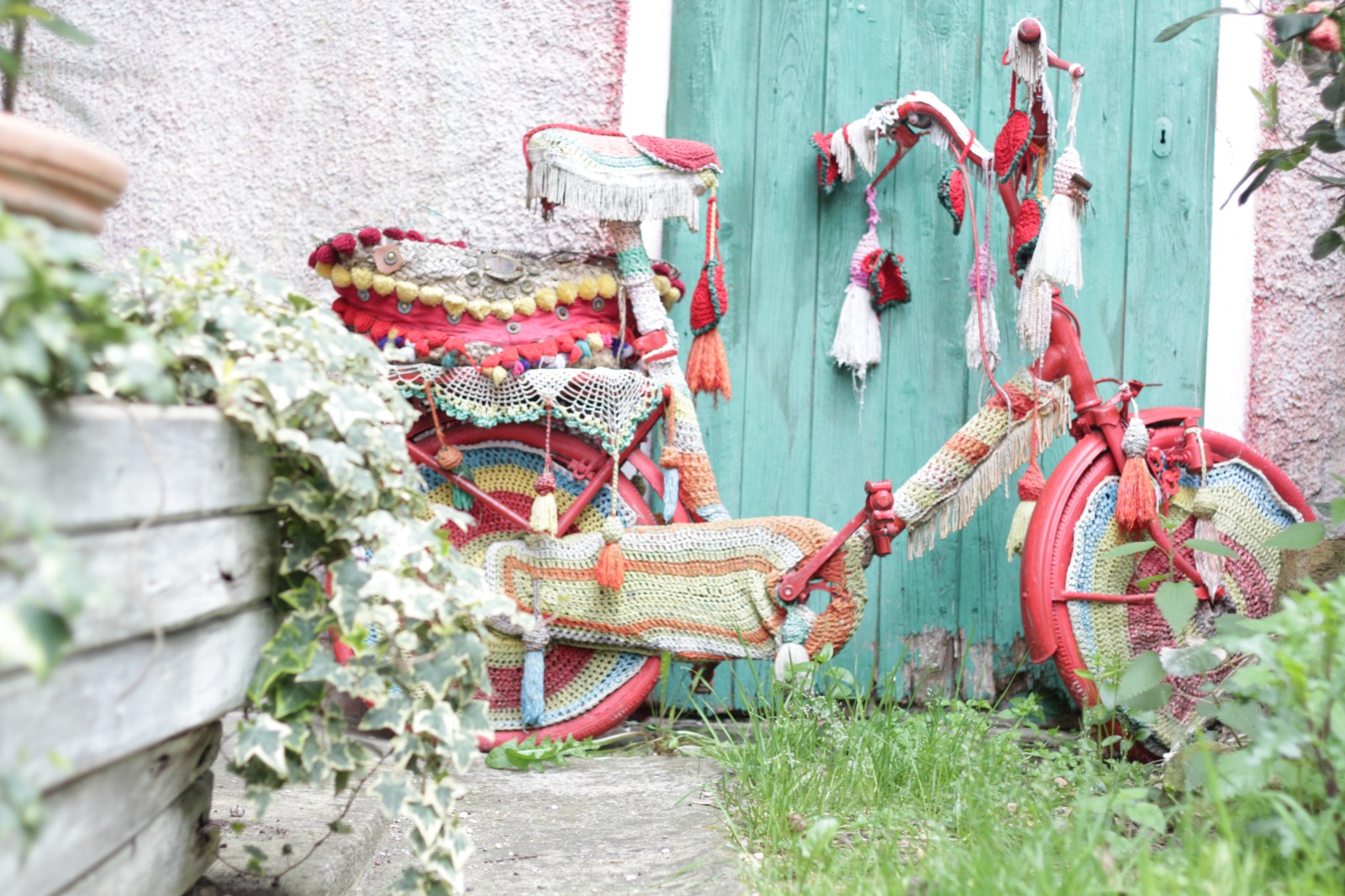 Fotografia di Rebecca Levi raffigurante una bicicletta decorata con tessuti colorati e lavorazioni all'uncinetto davanti a un portone verde.